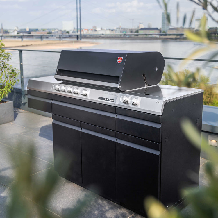 Otto-Wilde-G32 Gasgrill in Schwarz, stehend auf einer Terrasse, im Hintergrund ein großer Fluss mit Brücke und Teile einer Stadt. 